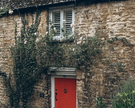 The rubble stone front of a cottage with a deep red panelled door, an iron knocker, a shuttered window above and climbing plants across the wall.