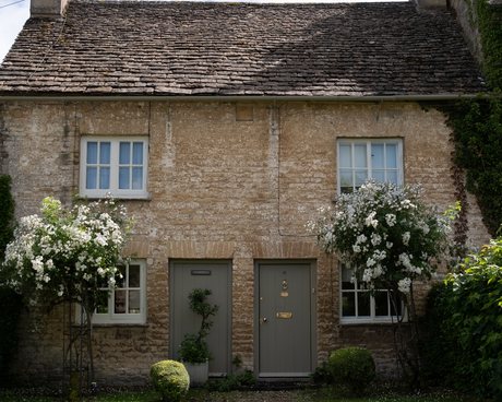 A pair of stone cottages under a stone-slate roof, with two painted front doors and white climbing roses trained around them.