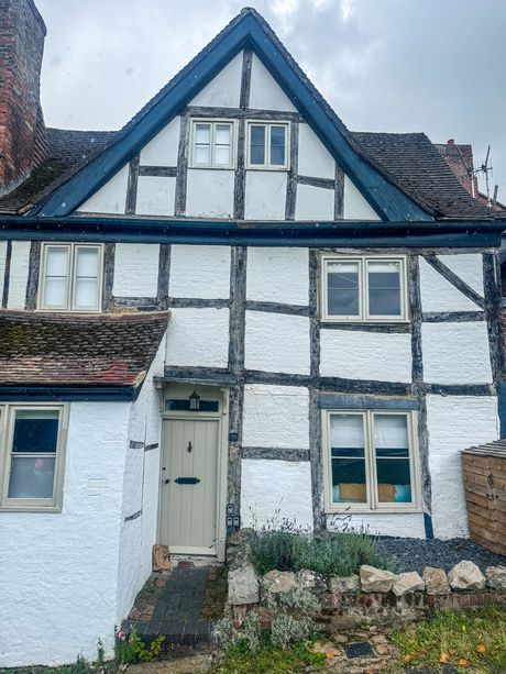 A white-painted timber-framed house with exposed dark timbers, a steep gable with two small windows, a tiled roof and a pale front door behind a low stone wall.